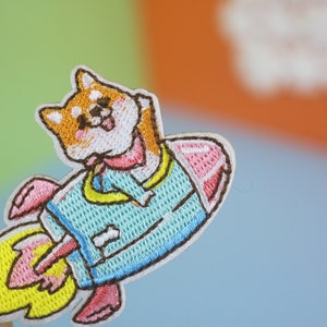 May include: Embroidered iron-on patch featuring a cartoon Shiba Inu dog wearing a blue spacesuit and riding a rocket with pink and yellow flames.