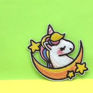 May include: Embroidered iron-on patch featuring a white unicorn with a pink and blue mane and tail, sleeping on a crescent moon with yellow stars.