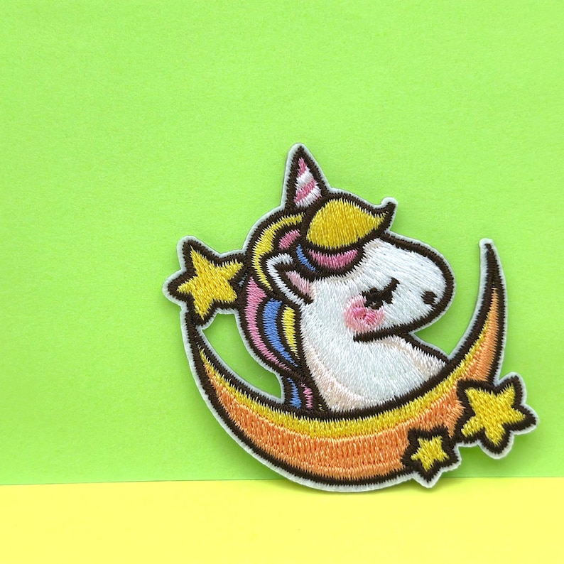 May include: Embroidered iron-on patch featuring a white unicorn with a pink and blue mane and tail, sleeping on a crescent moon with yellow stars.