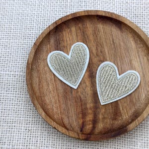 Puede incluir: Dos parches en forma de corazón con bordado de hilo dorado y borde azul claro se muestran sobre un posavasos redondo de madera. El posavasos tiene un color marrón intenso y un acabado liso. El fondo es una tela texturizada blanquecina.