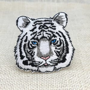 Puede incluir: Parche bordado con la cabeza de un tigre blanco con rayas negras, ojos azules y nariz rosa. El parche presenta costuras detalladas, perfecto para adornar prendas o accesorios. La cara del tigre está centrada en una tela texturizada de color blanquecino.