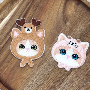 May include: Two embroidered patches featuring cat designs. One is a brown cat wearing a reindeer hat, the other is a pink cat with a hat. Both have detailed stitching and large eyes.