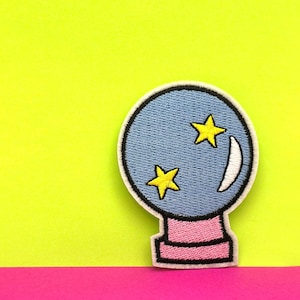 Op de afbeelding: Een geborduurde patch in blauw en roze met een kristallen bol met twee gele sterren erin. De kristallen bol staat op een roze voet.