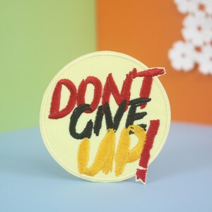 Könnte beinhalten: Gestickter Aufnäher mit den Worten "DON'T GIVE UP!" in roter, schwarzer und gelber Farbe auf einem hellgelben, kreisförmigen Hintergrund. Der Aufnäher hat einen weißen Rand und eine kleine rote Ausrufezeichen-Erweiterung.