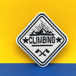 Puede incluir: Parche blanco en forma de diamante con texto bordado en negro que dice "CLIMBING" con dos estrellas a cada lado del texto. El parche también presenta una escena de montaña bordada en negro con un sol y dos hachas.