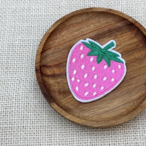 Puede incluir: Un parche de fresa rosa con detalles de semillas blancas y un tallo verde se encuentra en un plato pequeño y redondo de madera. El parche tiene un borde blanco. El plato es de madera oscura y está sobre un fondo neutro y texturizado.