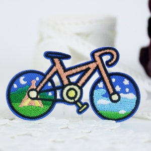 May include: An embroidered iron-on patch of a bicycle with a blue sky and clouds in one wheel and a green field with a tent and a crescent moon in the other wheel.