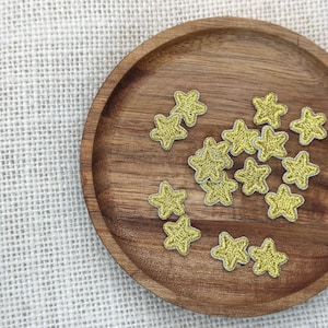 Puede incluir: Un conjunto de parches bordados con forma de estrella dorada sobre una bandeja redonda de madera. Los parches tienen un brillo metálico y están esparcidos por la bandeja. La bandeja está sobre un fondo de tela texturizada blanquecina.