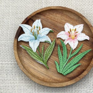 May include: Two embroidered lily flower patches, one light blue and one pink, with green leaves, rest on a round wooden plate. The flowers have detailed stitching and yellow stamens. The plate sits on a textured, off-white surface.