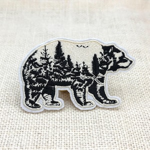 May include: Embroidered patch featuring a bear silhouette with a forest scene inside. The bear is black and white with a cream-colored border. The patch is likely for clothing or accessories.