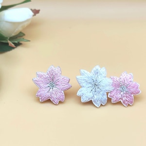 May include: Three embroidered flower patches in shades of pink and silver. The patches are in the shape of a flower with multiple petals. The background is a light peach color. A white flower and green leaves are in the upper left corner.