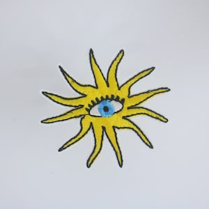 Könnte beinhalten: Eine gelbe gestickte Sonne mit einem blauen Auge in der Mitte. Die Sonne hat 12 Strahlen.