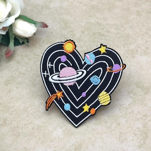 Puede incluir: Parche bordado en forma de corazón negro con un tema espacial. El diseño incluye planetas, estrellas y un sol en varios colores como rosa, amarillo, azul y morado. El parche es probablemente para ropa o accesorios.