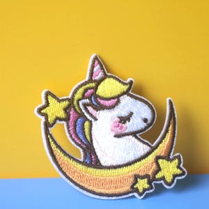 May include: A white unicorn with a rainbow mane and tail is sleeping on a yellow crescent moon with stars. The unicorn has a pink nose and closed eyes. The moon has a yellow and orange gradient.