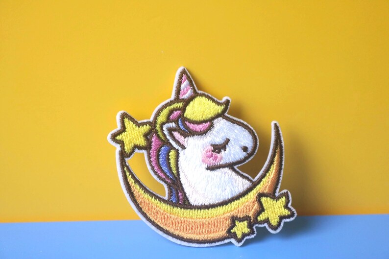 May include: A white unicorn with a rainbow mane and tail is sleeping on a yellow crescent moon with stars. The unicorn has a pink nose and closed eyes. The moon has a yellow and orange gradient.