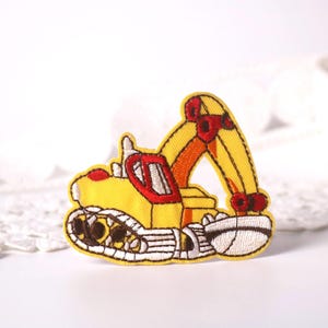 Könnte beinhalten: Gelber gestickter Bügelaufkleber mit einem Cartoon-Bagger mit roten Akzenten. Der Bagger hat einen gelben Körper, eine rote Kabine und einen gelben Arm mit einer roten Schaufel.