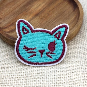 May include: An embroidered patch shaped like a cat head with bright turquoise thread for the face and dark maroon thread for the outline, ears, whiskers, and a winking eye. The patch features a white border edge and a flat, stitched texture.