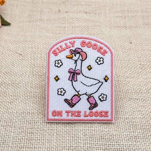 Puede incluir: Un parche bordado con fondo blanco y borde rosa. Presenta una oca blanca con un sombrero rosa, un lazo rosa y calentadores rosas. El texto SILLY GOOSE ON THE LOOSE está bordado en rosa. Pequeñas flores blancas y estrellas amarillas rodean a la oca.
