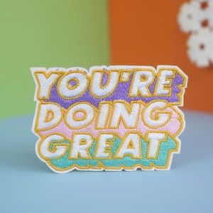 Może przedstawiać: Kolorowa naszywka haftowana z tekstem "YOU'RE DOING GREAT" w białych literach obwiedzionych żółtym kolorem. Naszywka znajduje się na jasnoniebieskim tle.