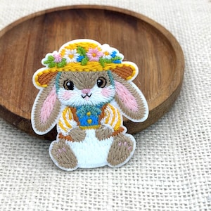 May include: An embroidered patch featuring a brown rabbit with pink inner ears, wearing a yellow straw hat adorned with pink, white, and blue flowers. The rabbit has a blue vest with yellow accents over a white shirt and is sitting in a forward-facing pose.