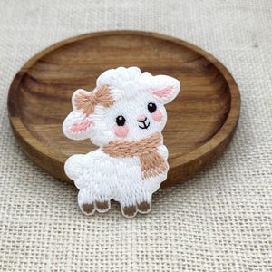 May include: An embroidered patch featuring a white lamb with a tan scarf and a matching tan bow on its head. The lamb has pink cheeks, black eyes, and brown hooves. The embroidery is detailed with white, tan, and pink thread on a white base.