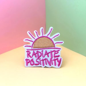 Peut inclure: Patch brodé rose avec un fond blanc représentant un soleil stylisé et le texte "RADIATE POSITIVITY".