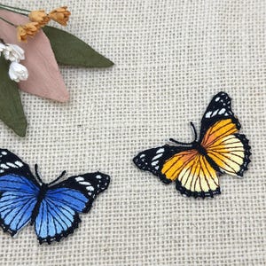 Puede incluir: Dos parches bordados de mariposas. Uno es azul con detalles negros, el otro es naranja y amarillo con detalles negros. Las mariposas están sobre un fondo beige texturizado, con un pequeño arreglo floral en la esquina superior izquierda.
