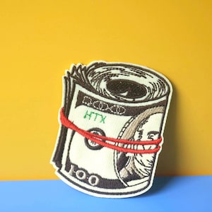 Puede incluir: Un parche bordado marrón y blanco de un rollo de dinero con una banda elástica roja. El parche tiene el texto "100" y "HTX" en él.