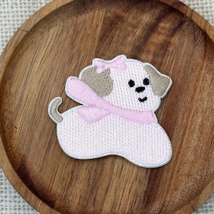 Puede incluir: Un parche bordado que muestra un cachorro de color rosa claro con orejas de color canela, un pequeño lazo rosa en la cabeza y una bufanda rosa alrededor del cuello. El cachorro tiene ojos y nariz bordados en negro, con un borde blanco.