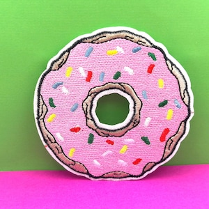 May include: A pink embroidered donut patch with colorful sprinkles. The donut is a popular food item and is often used as a decorative element on clothing, bags, and other accessories.