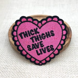 Könnte beinhalten: Ein pinkfarbener, herzförmiger, gestickter Aufnäher mit dem Text "THICK THIGHS SAVE LIVES" in Schwarz. Der Aufnäher hat einen gewellten schwarzen Rand und kleine Kreuzakzente. Der Aufnäher liegt auf einer Holzoberfläche.