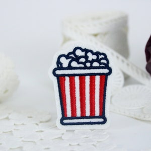 Pode incluir: Um balde de pipoca listrado vermelho e branco com contorno azul e pipoca branca dentro. O balde é bordado em um tecido branco.