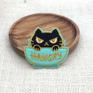 May include: Embroidered patch featuring a black cat with yellow eyes and whiskers, peeking over a turquoise bowl. The word "HANGRY" is embroidered in yellow. The patch is on a wooden plate.