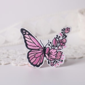 May include: Pink embroidered butterfly with floral details. The butterfly is outlined in black thread and has a pink and white floral design on one side.