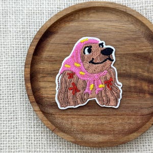 Puede incluir: Parche bordado con un perro de dibujos animados que lleva una bufanda y un gorro rosas con detalles amarillos. El perro es marrón con una nariz negra y una expresión sonriente. El parche está sobre una bandeja de madera.