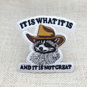 以下が含まれることがあります： カウボーイハットをかぶったアライグマの刺繍パッチ。「IT IS WHAT IT IS」の文字がアライグマの上に、「AND IT IS NOT GREAT」の文字が下にあります。アライグマは白黒で、茶色と黄色の帽子をかぶっています。