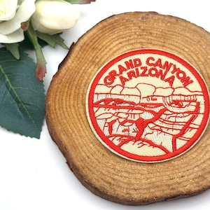 Op de afbeelding: Een ronde patch met de woorden "Grand Canyon Arizona" in het rood, en een rode illustratie van de canyon. De patch ligt op een houten schijf, met witte bloemen en groene bladeren op de achtergrond.