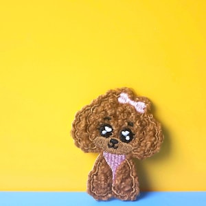 May include: A brown embroidered poodle with a pink bow and big eyes. The poodle is standing on a blue surface with a yellow background.