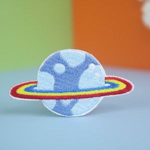 May include: A blue and white embroidered patch of a planet with a rainbow ring around it.