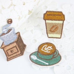 Op de afbeelding: Drie geborduurde strijk-op patches met koffiemotieven: een koffiemolen, een kop koffie en een koffiebeker om mee te nemen.