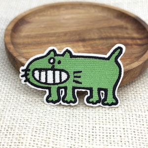 May include: A green embroidered cat patch with a white border. The cat has a large, toothy grin and is in a walking pose. The patch is on a light-colored fabric and a wooden plate.