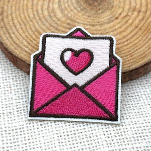 May include: Embroidered patch featuring a pink envelope with a white letter and a pink heart. The patch has a white border and is set against a wood slice background. The design is a symbol of love and romance.