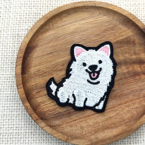 May include: Embroidered patch featuring a smiling white dog with pink ears and tongue, outlined in black. The patch is placed on a round, wooden plate with a natural wood grain finish. The dog has a fluffy appearance.