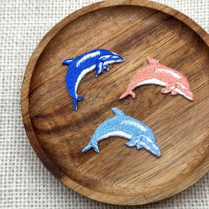 May include: Three embroidered dolphin patches in blue, light blue, and coral pink with white accents, arranged on a round wooden tray. The patches are detailed with stitched outlines and are set against a neutral linen background.