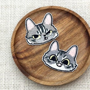 May include: Two embroidered cat face patches with gray and white details, set on a wooden plate. The cat faces have black eyes, pink ears, and white outlines. The patches are likely for clothing or accessories.