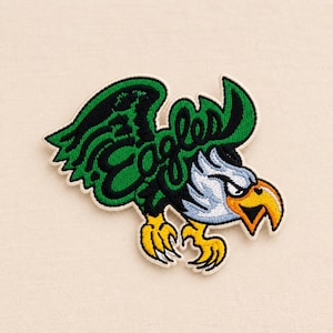 Könnte beinhalten: Ein gestickter Aufnäher mit einem stilisierten Adler mit weißem Kopf, gelbem Schnabel und grünen Flügeln. Das Wort Eagles ist in schwarzer Schreibschrift über den grünen Flügel gestickt. Das Design hat einen weißen Rand und detaillierte Stickerei.