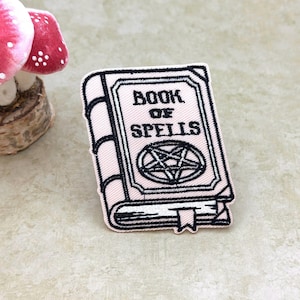 May include: A rectangular embroidered patch featuring a light pink book with black outlines and the text BOOK OF SPELLS printed on the cover above a black pentagram symbol. The patch includes a small bookmark detail at the bottom edge.