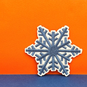 Puede incluir: Un parche bordado en forma de copo de nieve azul sobre un fondo naranja y azul.