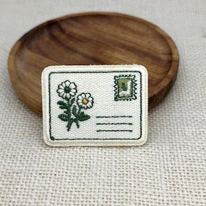 May include: A rectangular embroidered patch featuring an off-white envelope design with a green border. The patch displays two white daisies with yellow centers on the left and a small green postage stamp graphic in the upper right corner.