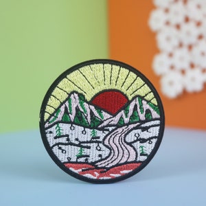 May include: A round embroidered patch with a mountain scene. The patch features a red sun setting behind a green mountain range with a white river winding through the valley. The background is a yellow sky with rays of light emanating from the sun.